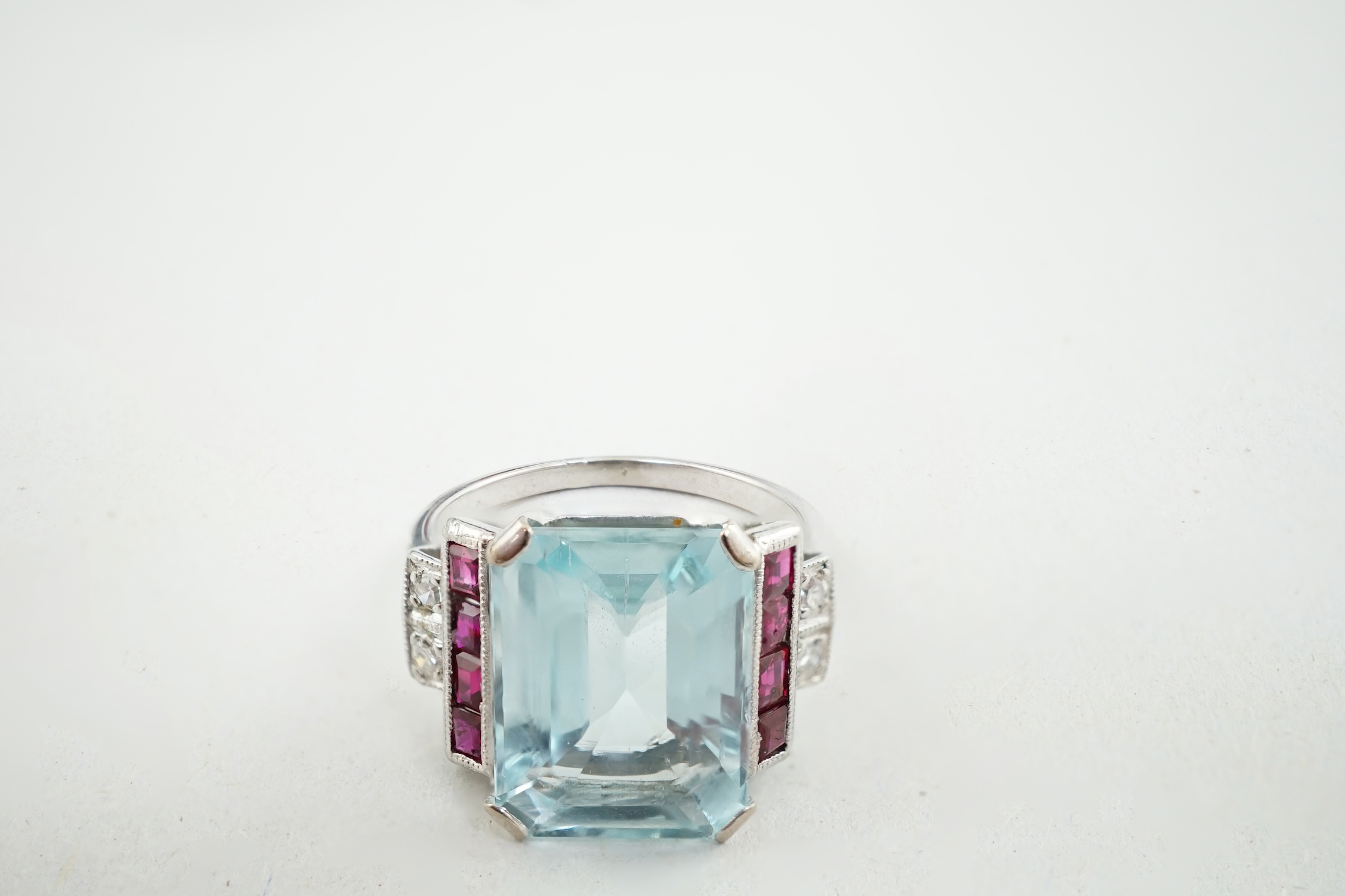 An Art Deco style platinum? and single stone emerald cut aquamarine set dress ring, with millegrain set ruby and diamond cluster stepped shoulders